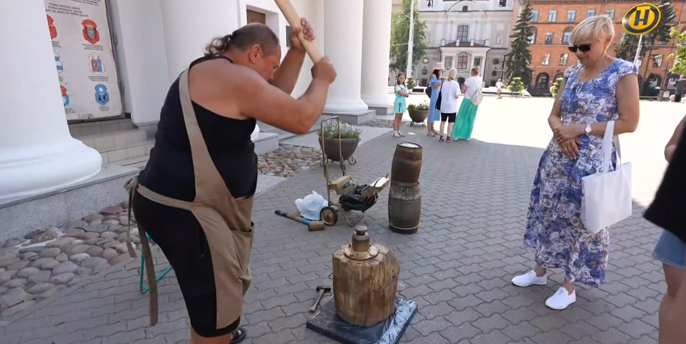 Кузнец в фартуке замахивается молотом над наковальней на деревянной колоде, чеканя монету на улице. Женщина в синем платье наблюдает за ним. Вверху справа желтый логотип ОНТ.