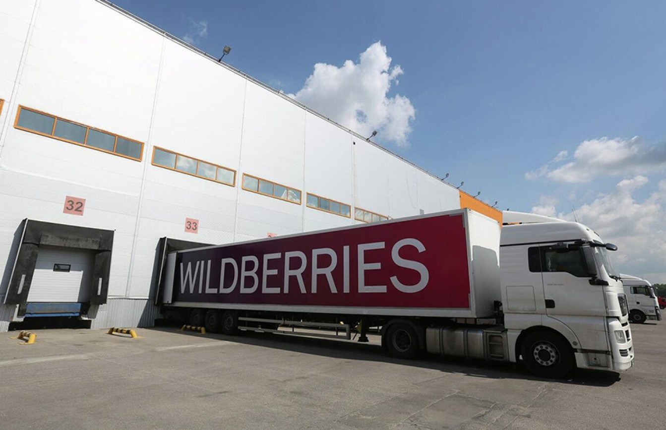 Грузовой автомобиль с логотипом Wildberries припаркован у погрузочного шлюза крупного современного складского комплекса под ясным небом с редкими облаками.