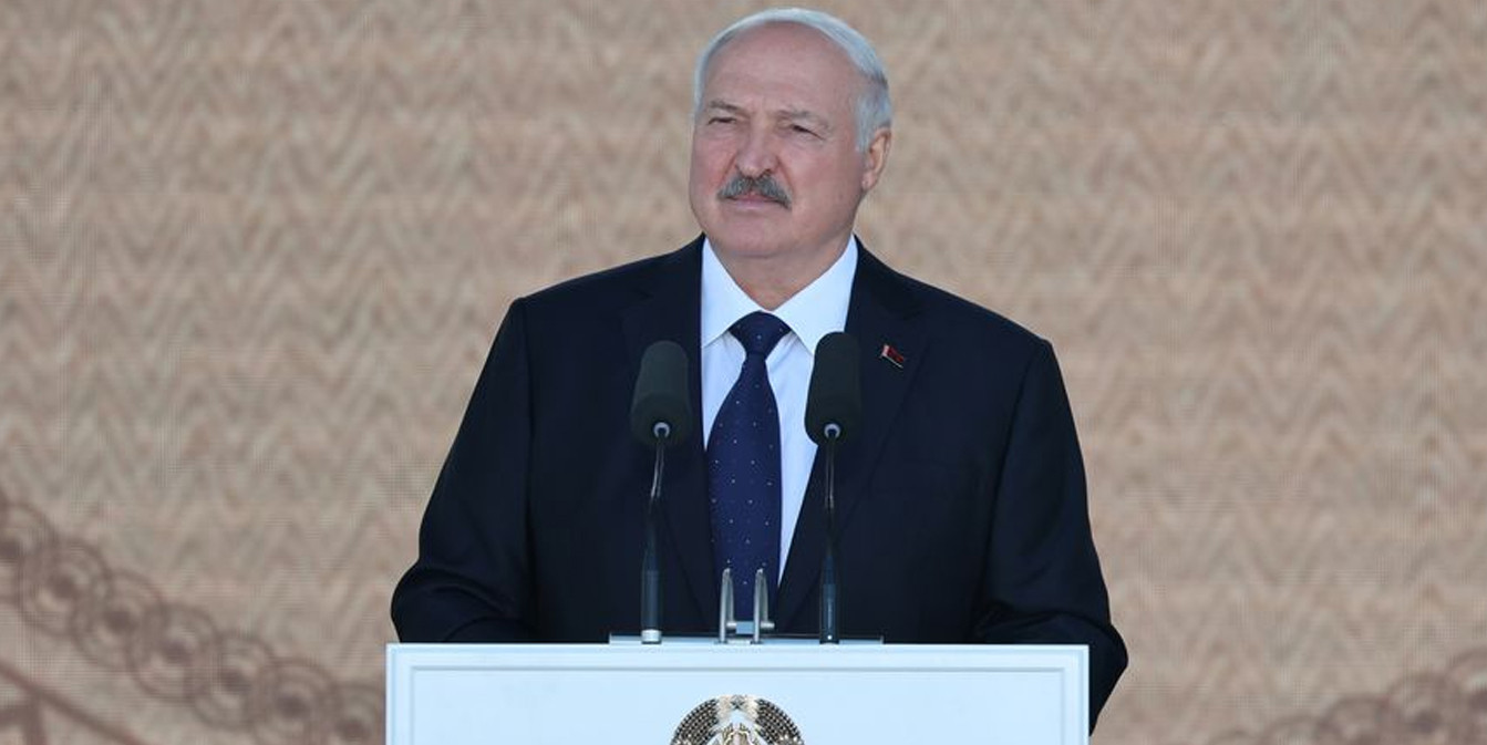 Президент Александр Лукашенко выступает за трибуной на сцене фестиваля «Зов Полесья» («Кліч Палесся»). Сзади огромные экраны с народным орнаментом. Перед сценой сидят зрители.