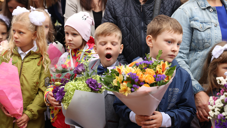 Группа маленьких детей стоит на школьной линейке в окружении взрослых. На переднем плане мальчик в сером пиджаке с бабочкой широко и заразительно зевает, закрыв глаза.